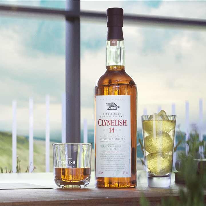 A bottle of Clynelish 14 Year Old Single Malt Whisky stands on a table between a whisky glass and an ice-filled glass, set against a scenic outdoor backdrop that reflects its coastal character.