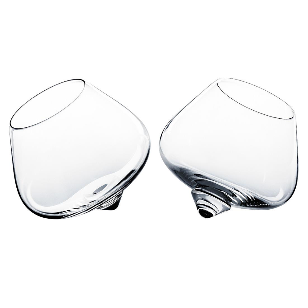 The Normann Copenhagen Cognac Glass 2 pcs features two clear, tilted glasses with rounded bottoms that gently rock without spilling, embodying contemporary Scandinavian design elegance against a white background.