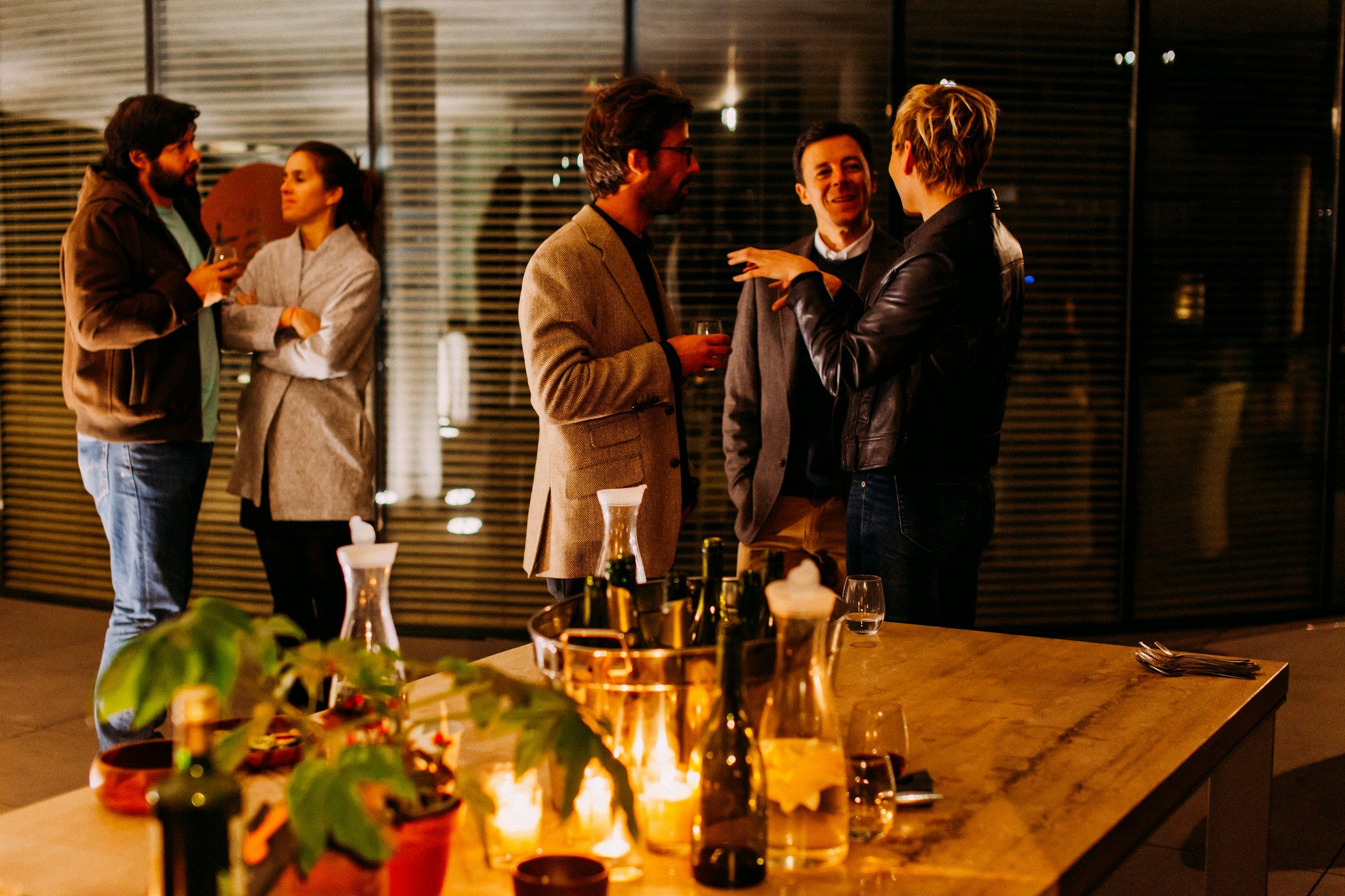 Group of people socializing around a table with drinks and plants in an indoor setting.