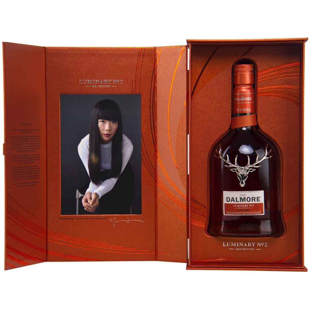 A gift box includes The Dalmore Luminary No. 2 16 Year Old Single Malt Scotch Whisky and a portrait photo of a woman with long black hair in a white and black outfit, set against an orange patterned background.