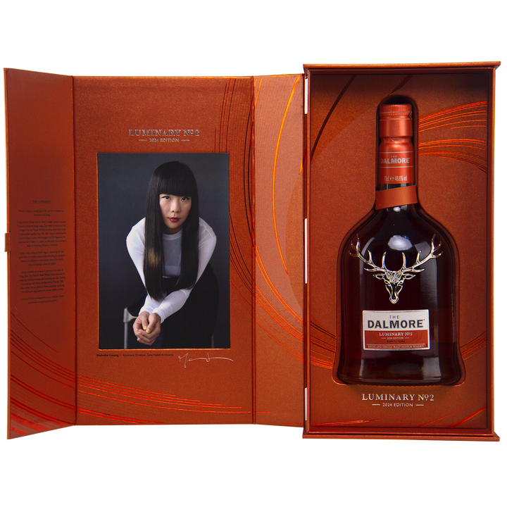 A gift box includes The Dalmore Luminary No. 2 16 Year Old Single Malt Scotch Whisky and a portrait photo of a woman with long black hair in a white and black outfit, set against an orange patterned background.