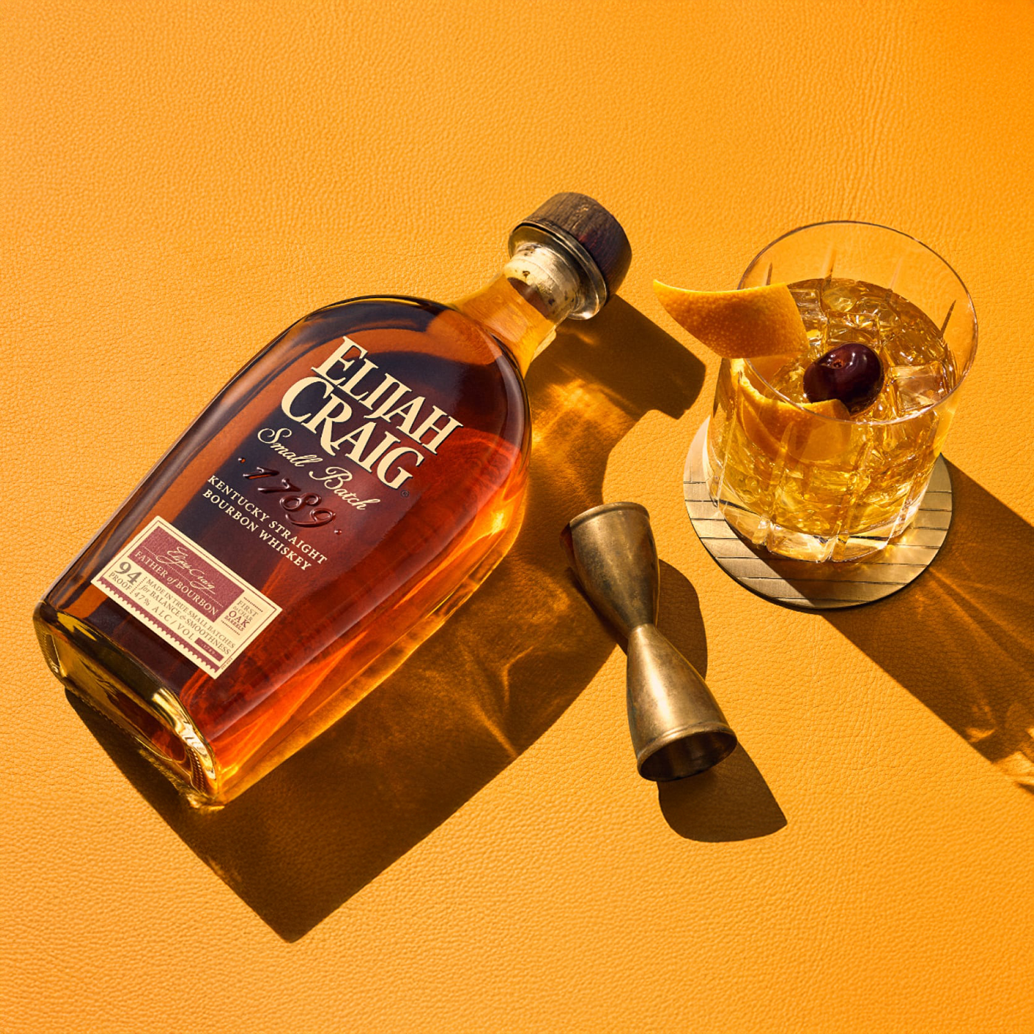 A bottle of Elijah Craig Small Batch Kentucky Straight Bourbon Whiskey rests on an orange surface next to a metal jigger and a glass of whiskey, garnished with an orange peel and cherry, all casting long shadows.