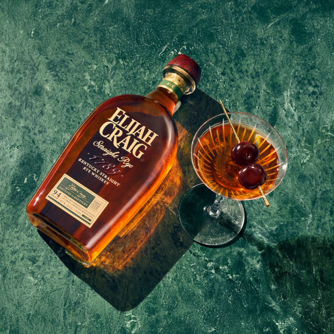A bottle of Elijah Craig Kentucky Straight Rye Whiskey sits on a green textured surface next to a cocktail glass filled with the same whiskey, garnished with two cherries on a skewer.