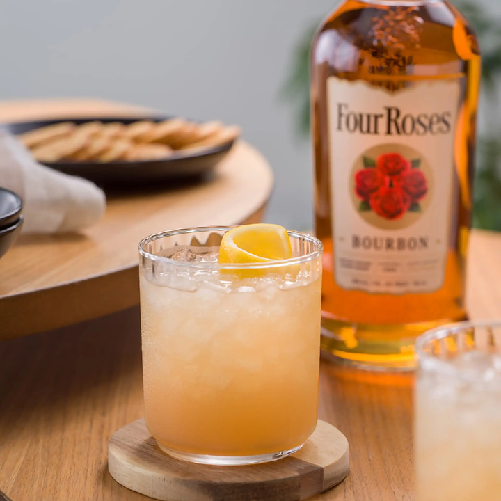 A glass of iced cocktail with a lemon twist rests on a wooden coaster, with a bottle of Four Roses Kentucky Straight Bourbon Whiskey and a plate of crackers in the background.