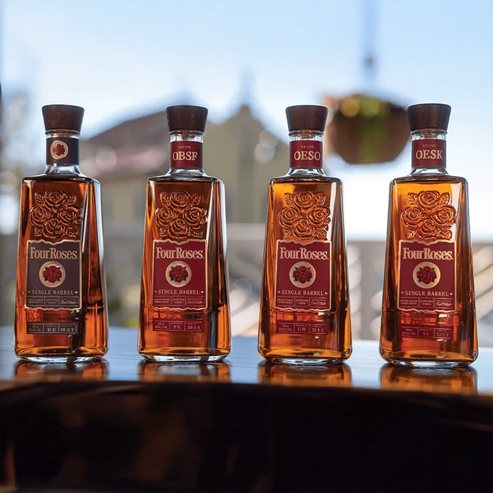 Four bottles of Four Roses Single Barrel Bourbon Whiskey, each 100 proof, stand in a row outdoors on a reflective surface with their unique recipe codes visible beneath the clear sky.