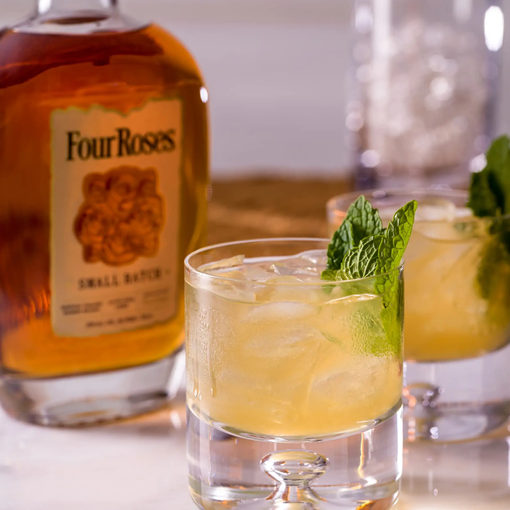 A glass of iced cocktail garnished with fresh mint sits before a bottle of Four Roses Small Batch Kentucky Straight Bourbon Whiskey, known for enhancing cocktails, with another similar drink and a glass jar in the background.