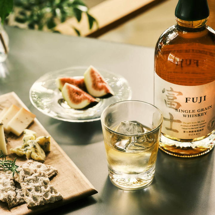 A glass of whiskey with ice sits beside a bottle of Fuji Single Grain Japanese Whisky, a plate of sliced figs, and a wooden board topped with assorted cheeses and crackers on the table.