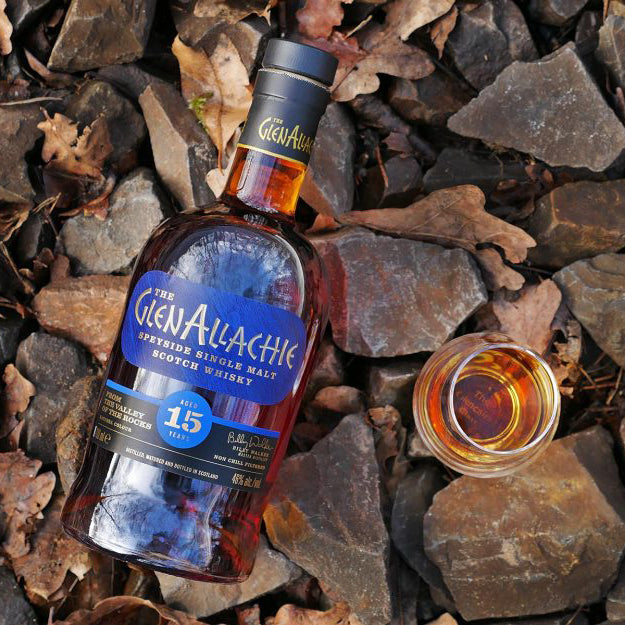 A bottle of GlenAllachie 15 Year Old Single Malt Scotch Whisky rests on rocks and leaves, beside a glass filled with this sherry cask matured whisky.