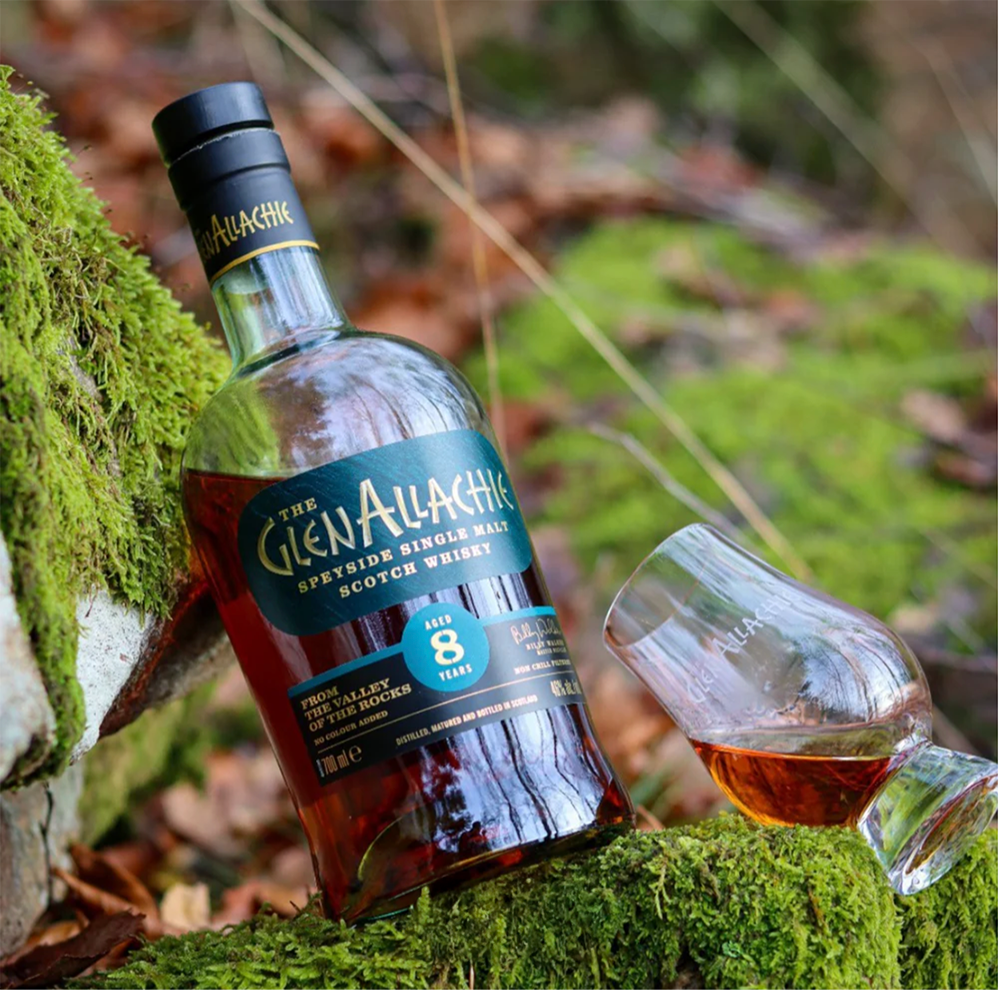 A bottle of GlenAllachie 8 Year Old Single Malt Scotch Whisky and a whisky glass sit on a mossy rock outdoors among fallen leaves.