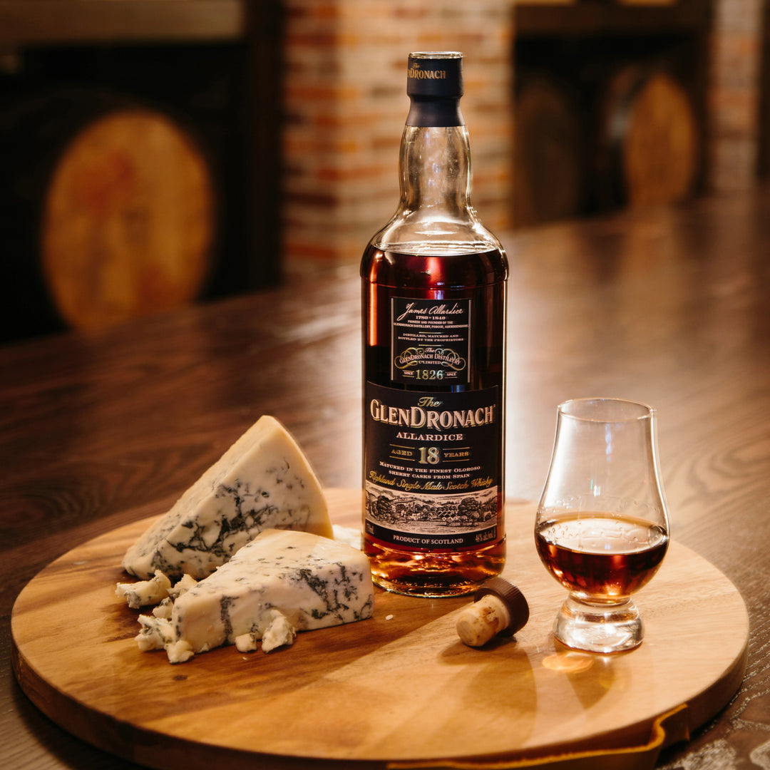 A bottle of Glendronach 18 Year Old Allardice Single Malt Scotch Whisky sits next to a glass of whisky and blue cheese slices on a wooden board, all arranged on a polished table with a rustic, blurred background.