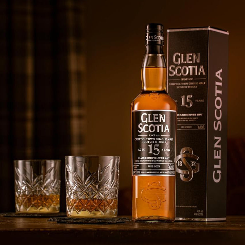 A bottle of Glen Scotia 15 Year Old Single Malt Scotch Whisky stands next to its box and two crystal glasses filled with whisky on a wooden table, warmly lit for a cozy, inviting atmosphere.