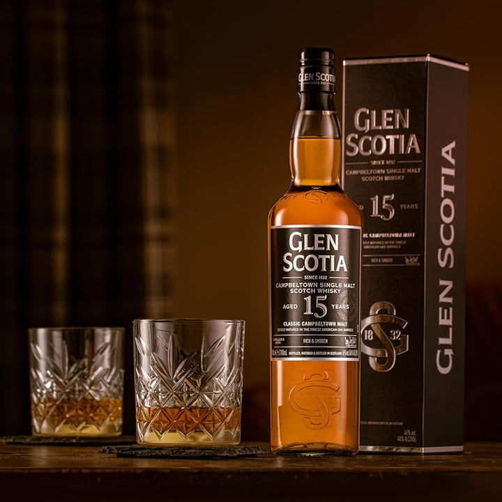 A bottle of Glen Scotia 15 Year Old Single Malt Scotch Whisky stands next to its box and two crystal glasses filled with whisky on a wooden table, warmly lit for a cozy, inviting atmosphere.