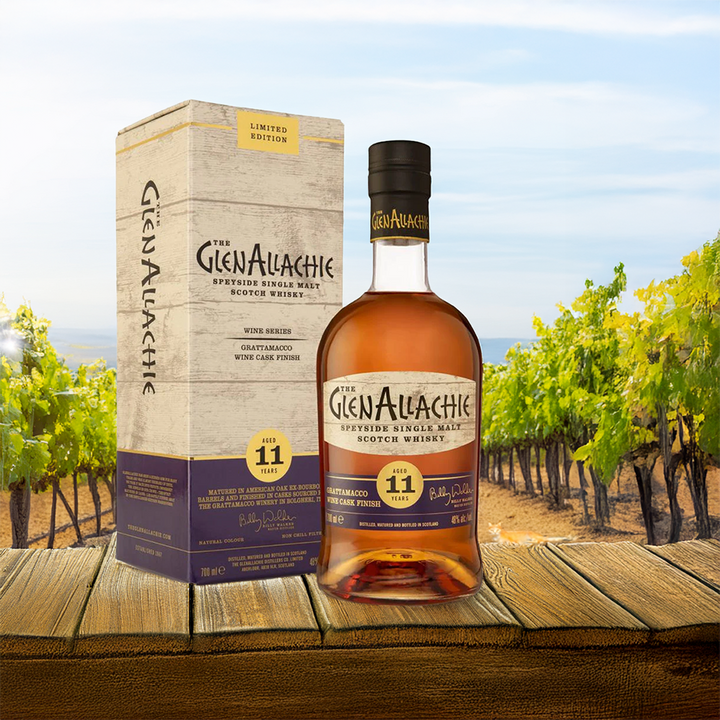 A bottle and box of GlenAllachie 11 Year Old Grattamacco Cask Finish Single Malt Scotch Whisky rest on a wooden table, with a vineyard in the background under a clear blue sky.