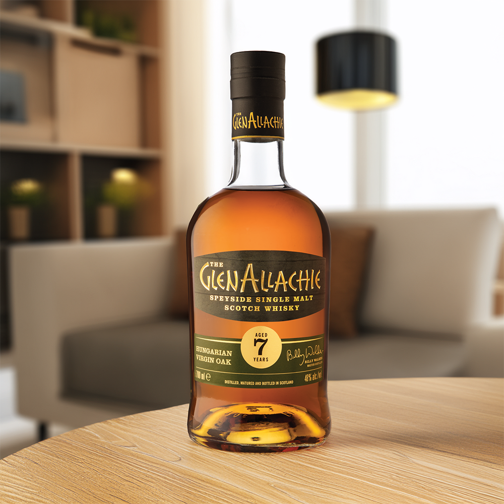 A bottle of GlenAllachie 7 Year Old Hungarian Oak Single Malt Scotch Whisky rests on a wooden table in a modern living room, with a sofa and lamp behind it, showcasing its special maturation in Hungarian oak casks.