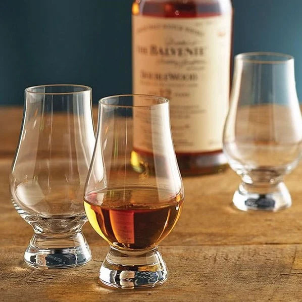 Three Glencairn Crystal Whisky Glasses (Black & White Box), one filled with amber whisky, rest on a wooden surface. A blurred bottle of Balvenie in the background against a blue wall sets the mood for an elegant whisky tasting.