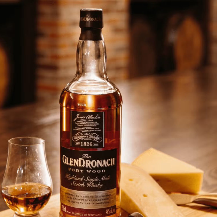 A bottle of Glendronach Port Wood Single Malt Scotch Whisky rests on a wooden table next to a glass of whisky and cheese slices, with a brick fireplace glowing warmly in the background—an inviting scene for this port cask finish.