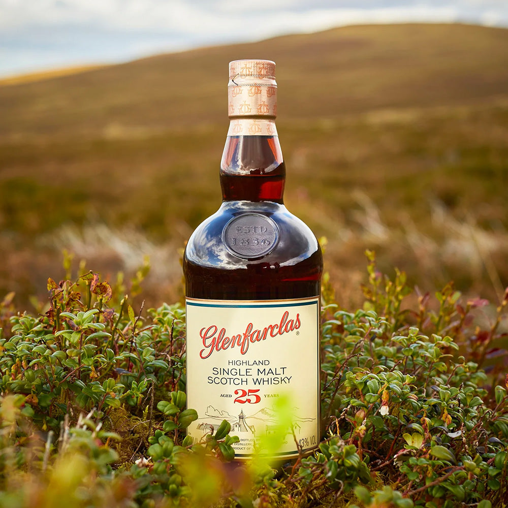 A bottle of Glenfarclas 25 Year Old Single Malt Scotch Whisky stands upright amid green shrubbery, its Oloroso sherry cask character reflected in the soft-focus hilly landscape behind.
