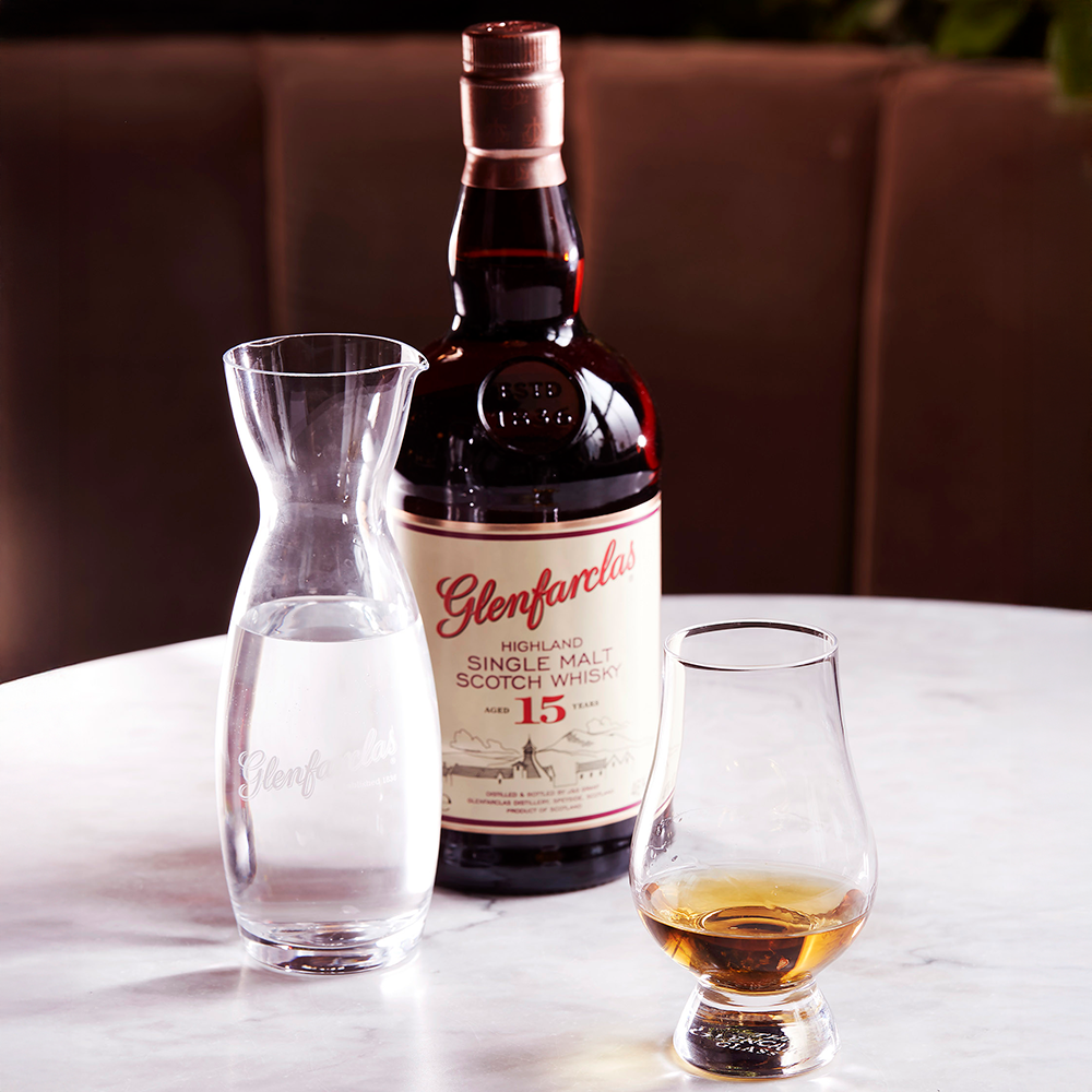 A bottle of Glenfarclas 15 Year Old Single Malt Scotch Whisky sits on a marble table, accompanied by a carafe of water and a glass containing a small pour of the single malt.
