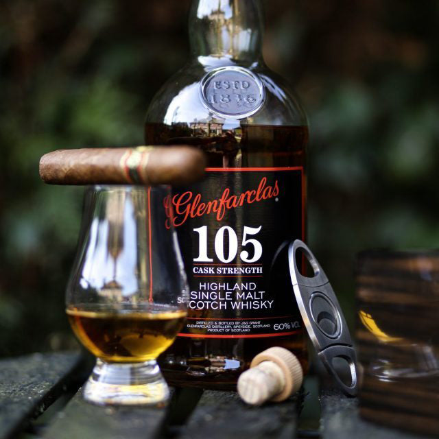 A bottle of Glenfarclas 105 Cask Strength Single Malt Scotch Whisky sits behind a whisky glass filled with Speyside single malt, with a cigar on top. A cigar cutter, wooden box, and bottle cork rest nearby on a wooden outdoor surface.