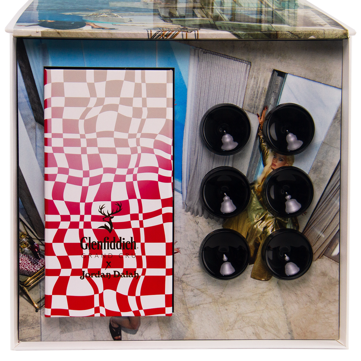 A Glenfiddich Grand Cru 23 Year Old x Jordan Dalah Box Set displays a red and white wavy checkered design, the brand logo, six black holders, and a woman in a gold dress standing in a modern room as the backdrop.