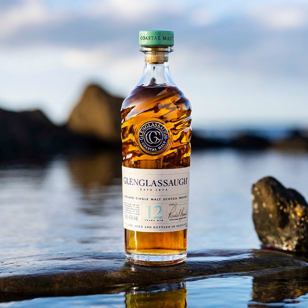 A bottle of Glenglassaugh 12 Year Old Single Malt Scotch Whisky stands on a wet rock by the sea, with waves crashing and blurred rocks in the background beneath a cloudy sky.