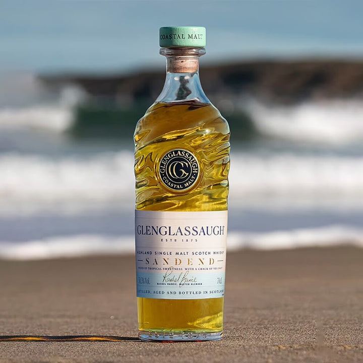 A bottle of Glenglassaugh Sandend Single Malt Scotch Whisky stands on a sandy beach with blurred ocean waves and rocks behind it. Its wavy design and blue-green label evoke the spirit of Scottish Highland whisky.