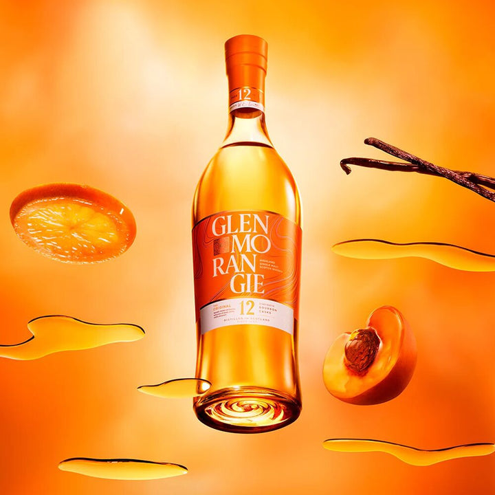 A bottle of Glenmorangie The Original 12 Year Old Single Malt Scotch Whisky sits among peach slices, a vanilla bean, and golden liquid splashes on a vivid orange background.