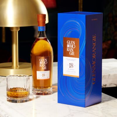 A bottle of Glenmorangie 18 Year Old Extremely Rare Single Malt Scotch Whisky stands by its blue gift box on a marble table, accompanied by a glass of the whisky and a gold lamp.
