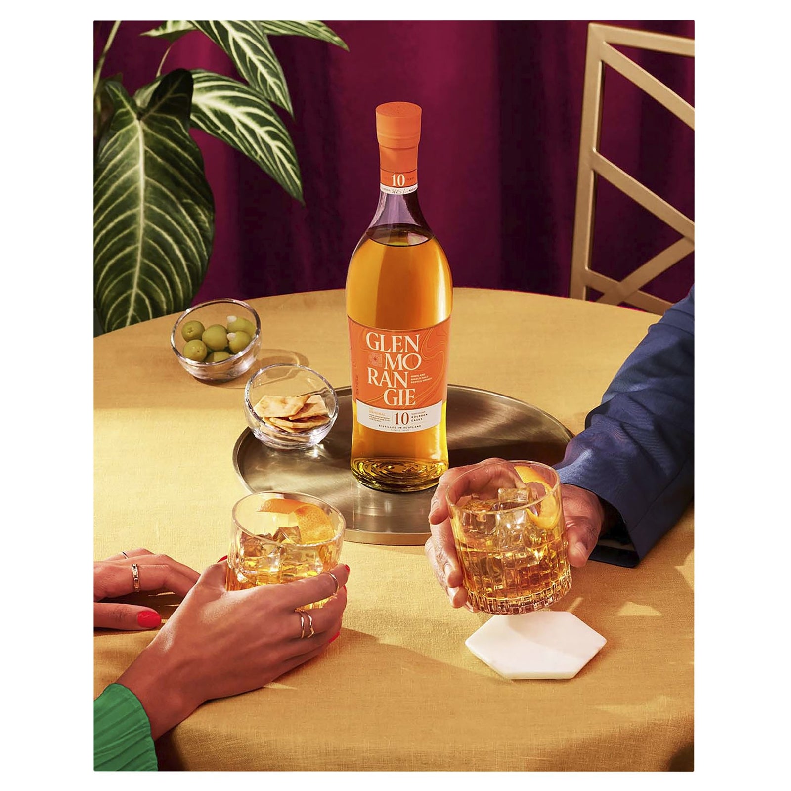A bottle of Glenmorangie 10 Year Old Single Malt Scotch Whisky is on a round tray with two whisky glasses, green olives, and crackers. Two people raise their glasses together over a yellow tablecloth.