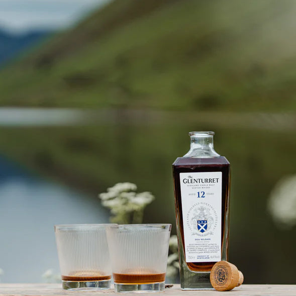 A bottle of Glenturret 12 Year Old Single Malt Scotch Whisky stands beside two filled glasses and a cork on a table, against a blurred backdrop of greenery and shimmering water.