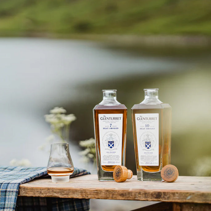 A bottle of Glenturret 7 Year Old Peat Smoked Single Malt Scotch Whisky sits on a wooden table with a whisky glass, corks, a blue tartan cloth, and a scenic lake with green hills in the background.
