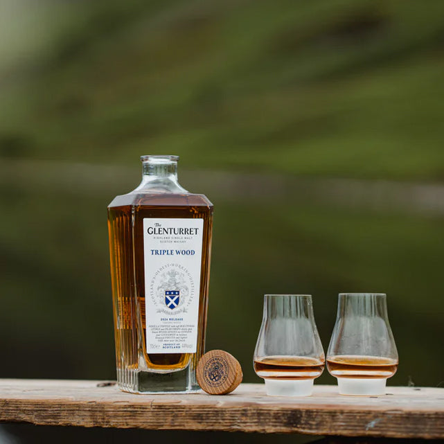 A bottle of Glenturret Triple Wood Single Malt Scotch Whisky stands on a wooden surface with its cork and two glasses of whisky, set against a blurred green background.