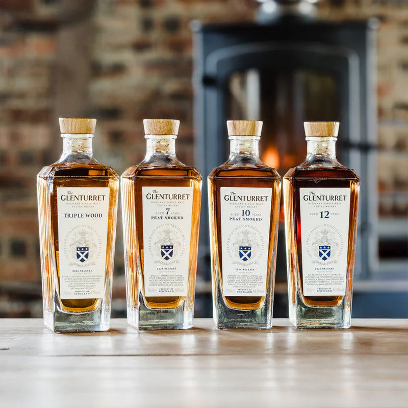 Four bottles of Glenturret single malt whisky, including Glenturret 10 Year Old Peat Smoked Single Malt Scotch Whisky, are displayed on a wooden table with a blurred fireplace and brick wall creating a warm backdrop.