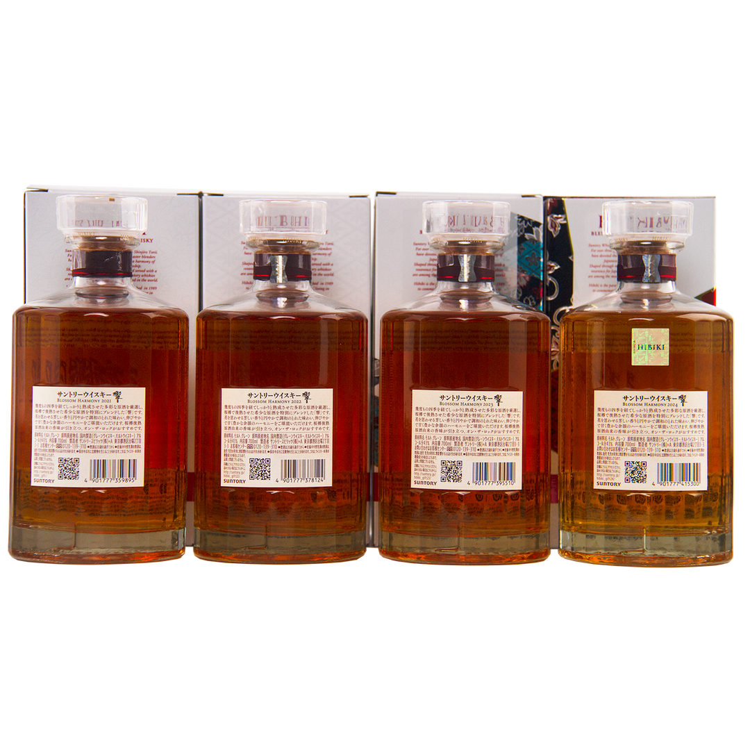 Four bottles from the Hibiki Blossom Harmony Limited Edition Collection 2021 to 2024 are lined up side by side, displaying amber liquid with clear faceted caps and back labels; boxes with Japanese text appear behind each bottle.