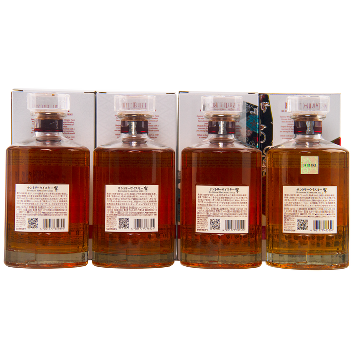 Four bottles from the Hibiki Blossom Harmony Limited Edition Collection 2021 to 2024 are lined up side by side, displaying amber liquid with clear faceted caps and back labels; boxes with Japanese text appear behind each bottle.