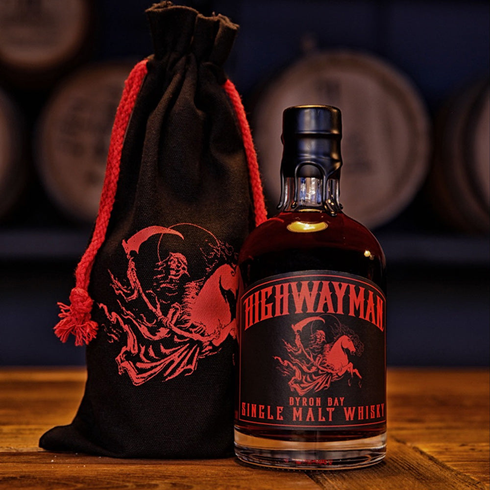 A bottle of Highwayman Far Beyond Peatera Single Malt Australian Whisky with a black and red skeleton label is displayed beside a matching black drawstring bag on wood, with blurred barrels in the background.