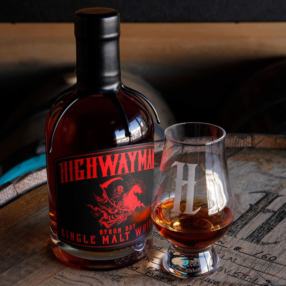 A bottle of Highwayman The Indiana Gentleman Single Malt Australian Whisky with a red and black label stands next to a whisky glass filled with cask strength amber liquid, both set on a wooden barrel.