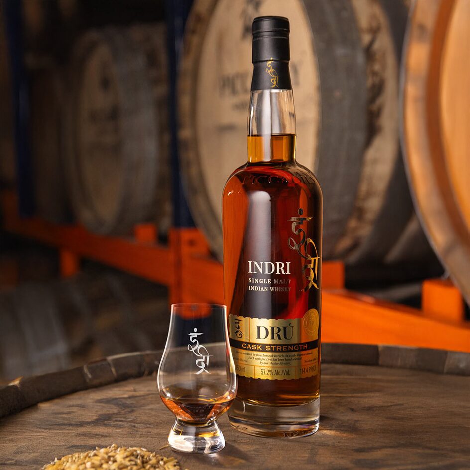 A bottle of Indri Drú Cask Strength Single Malt Indian Whisky sits next to a filled glass on a wooden barrel, surrounded by scattered grain, with blurred whisky barrels in the background.