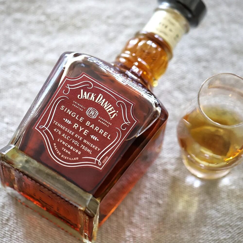 A bottle of Jack Daniel's Single Barrel 4 Year Old Rye Whiskey rests on its side beside a glass, displaying the rich amber liquid on a light textured surface.