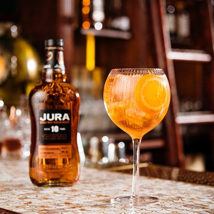 A bottle of Jura 10 Year Old Single Malt Scotch Whisky sits beside a large glass with an orange cocktail and orange slices on a patterned bar counter, with a blurred bar background.