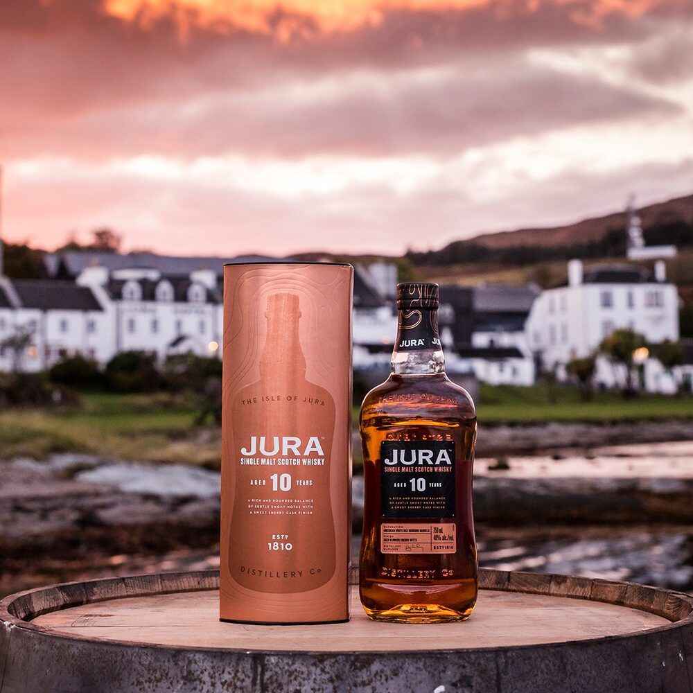 A bottle of Jura 10 Year Old Single Malt Scotch Whisky and its packaging rest on a barrel outdoors, framed by white buildings and a pink sunset, capturing the distinctive character of Jura Distillery.