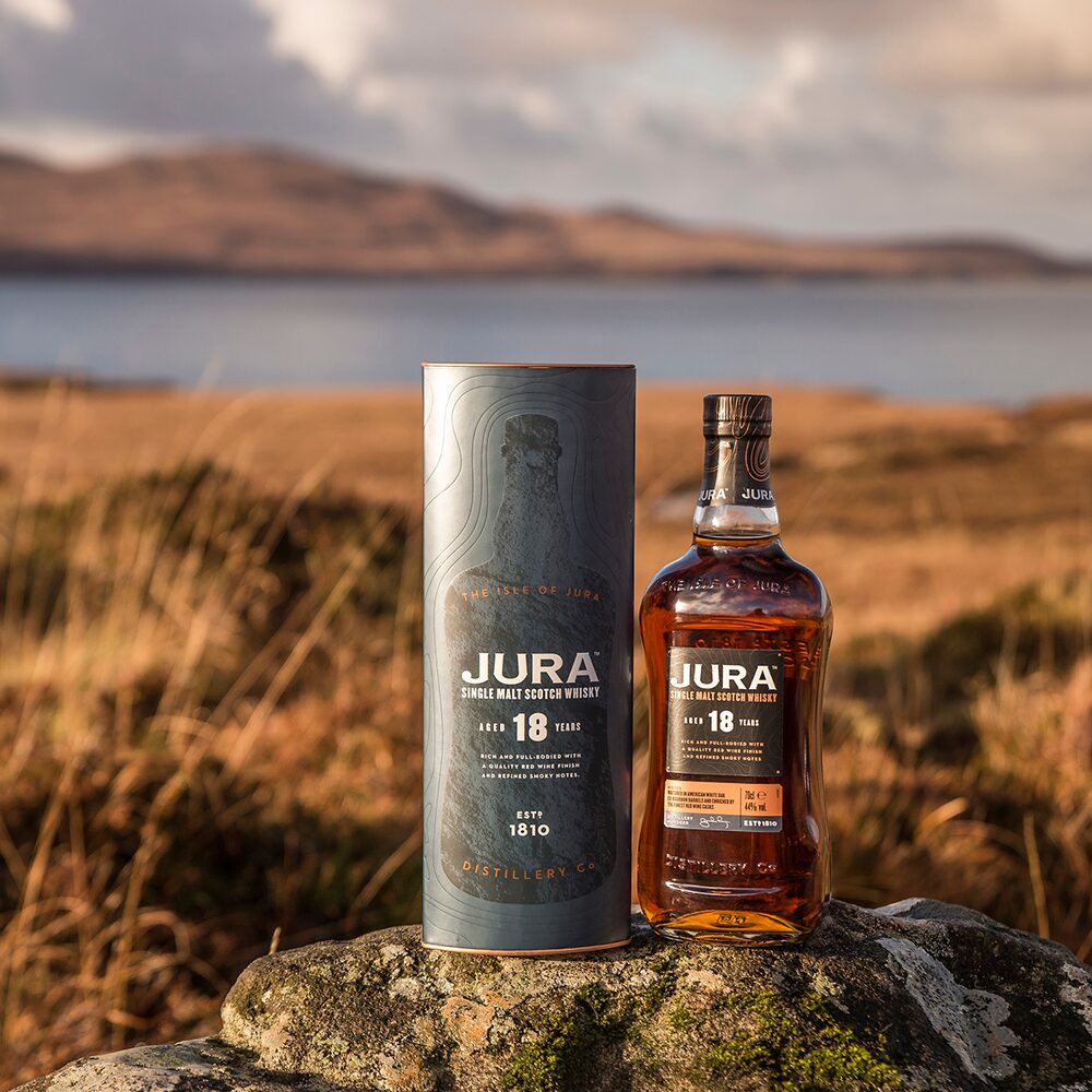 A bottle of Jura 18 Year Old Single Malt Scotch Whisky with its cylindrical box is placed on a rock outdoors, with hills, water, and grass in the blurred background—highlighting this whisky’s Bordeaux cask influence.