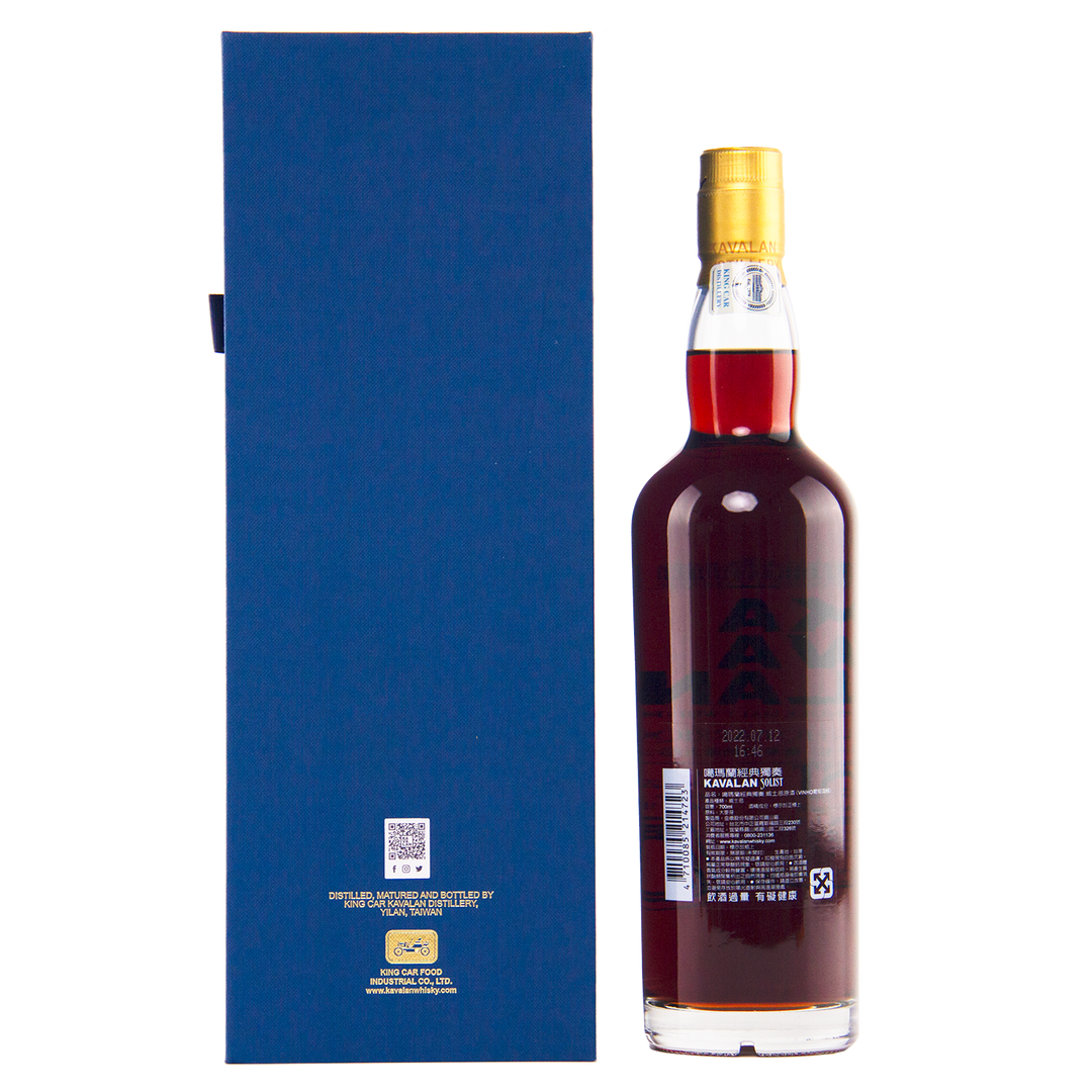 A bottle of dark amber Kavalan 2015 Vinho Barrique Cask No. W151217034A whisky with a gold cap stands beside a tall blue rectangular box displaying text and a QR code, both set against a plain white background.