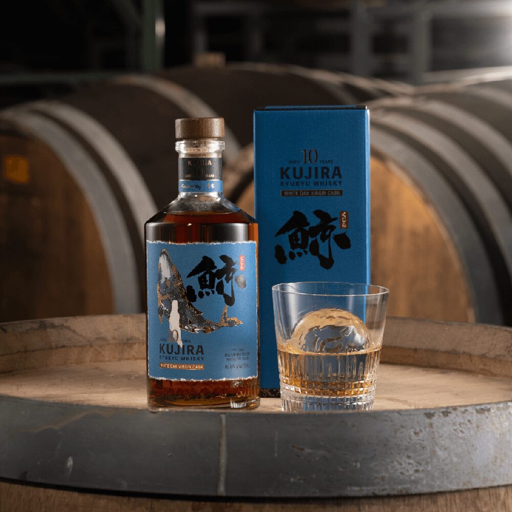 A bottle of Kujira Ryukyu 10 Year Old Japanese Whisky with its blue box and a whiskey glass filled with ice sits on a barrel in a dimly lit distillery, surrounded by large wooden casks in the background.