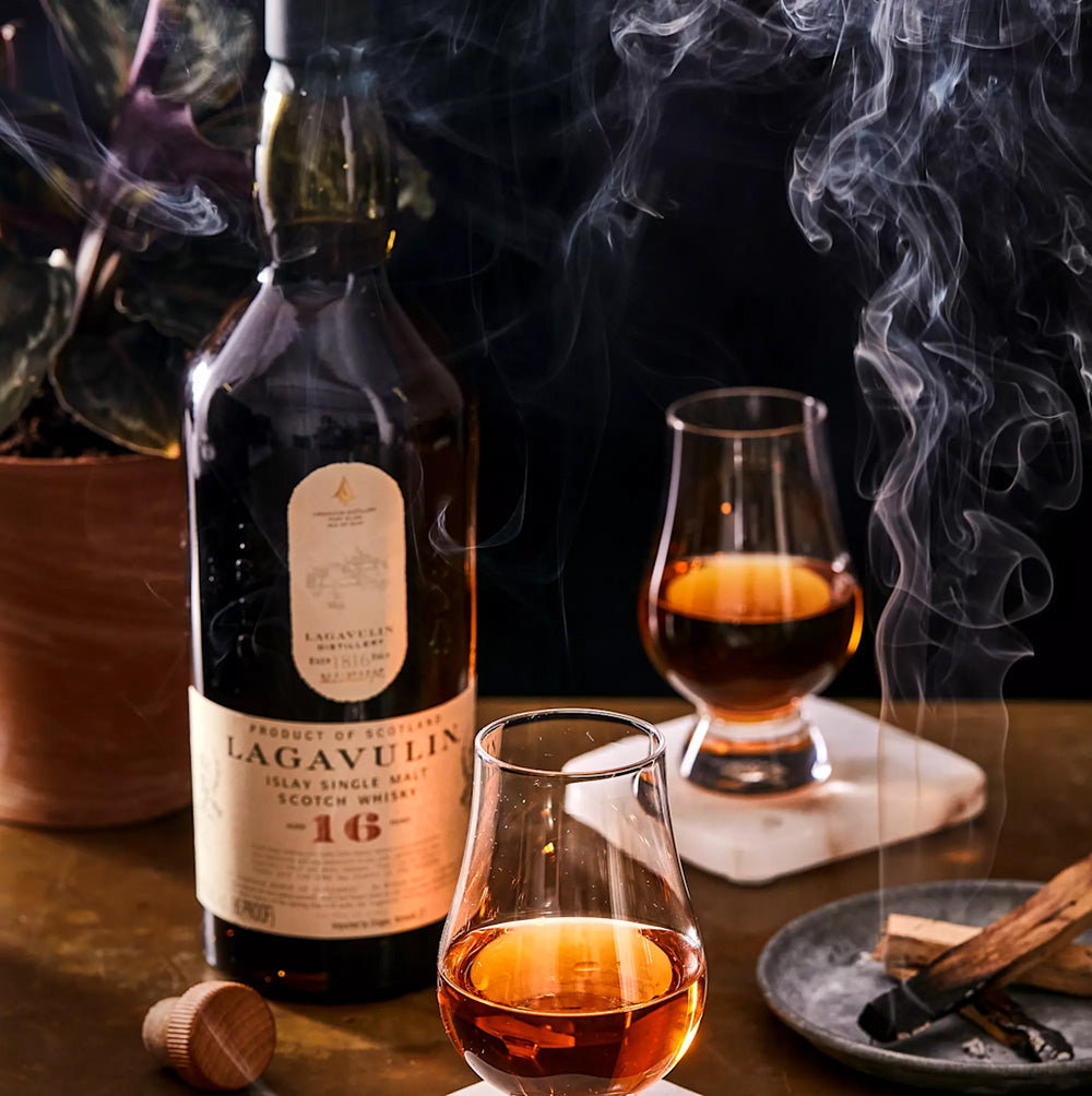 A bottle of Lagavulin 16 Year Old Single Malt Scotch Whisky stands on a table with two poured glasses, smoke swirling above a plant in the background. A whisky cork and a dish of charred wood sticks enhance this peated whisky scene.