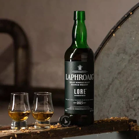 A bottle of Laphroaig Lore Single Malt Scotch Whisky stands beside two filled glasses on a wooden surface, with a rustic backdrop and scattered barley grains.