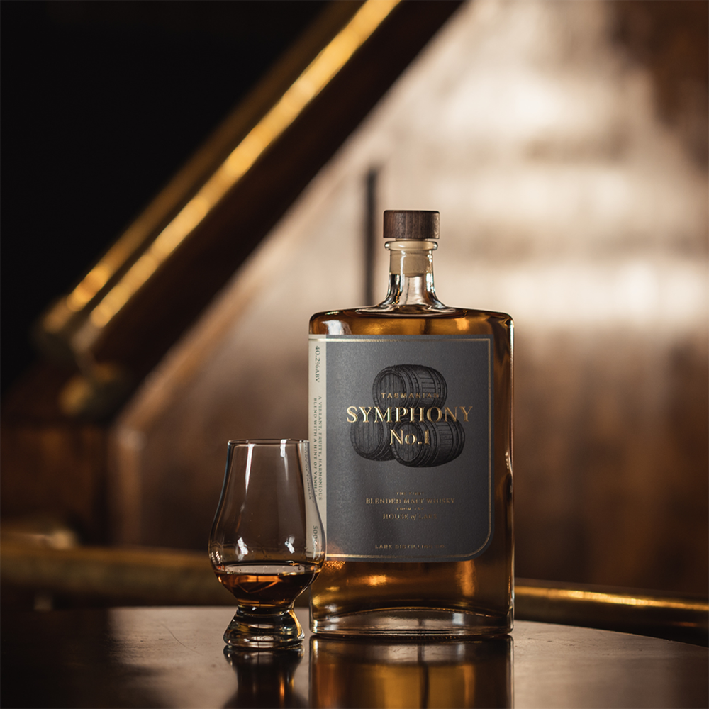 A bottle of Lark Symphony no.1 Tasmanian Whisky with a dark label and wooden cap stands next to a glass of whisky on a polished wooden surface, set against a softly blurred, warm-toned background.