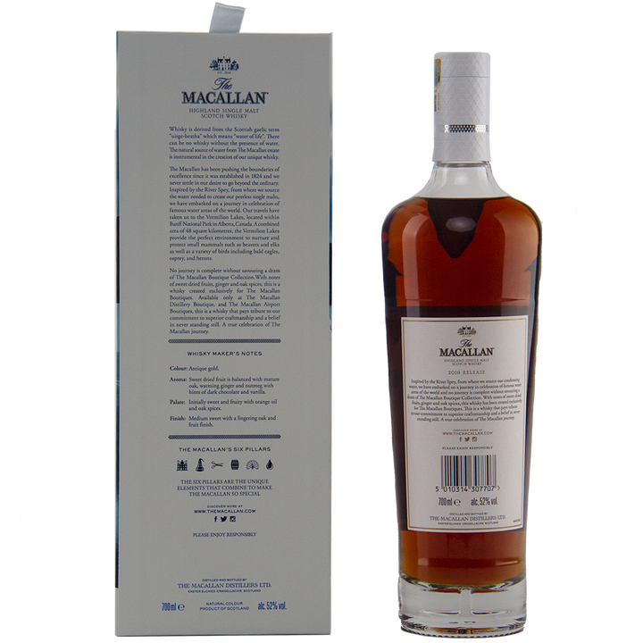 A bottle of The Macallan Boutique Collection 2019 Release Single Malt Whisky stands beside its grey box packaging. The dark amber whisky is presented in a white label with gold and black accents, highlighting its travel retail exclusivity.