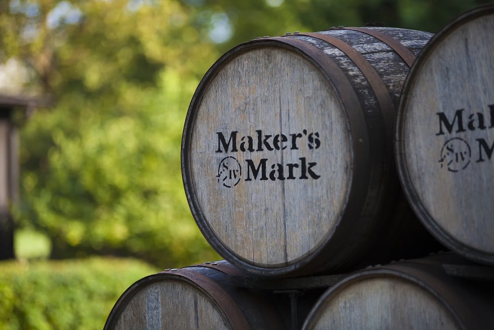 Wooden whiskey barrels stacked outdoors, with the barrel in focus showing the printed brand name 'Maker's Mark' on the stave, against a blurred green garden background.