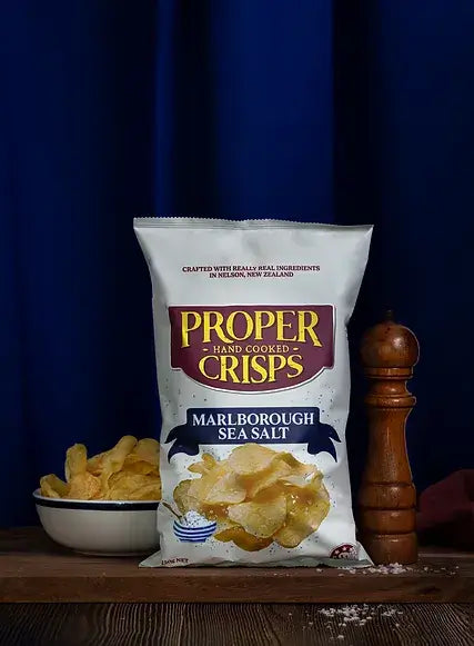 A bag of Proper Crisps Marlborough Sea Salt gluten-free crisps stands upright on a wooden surface beside a pepper grinder, a bowl of chips, and sea salt, all set against a dark blue curtain backdrop.